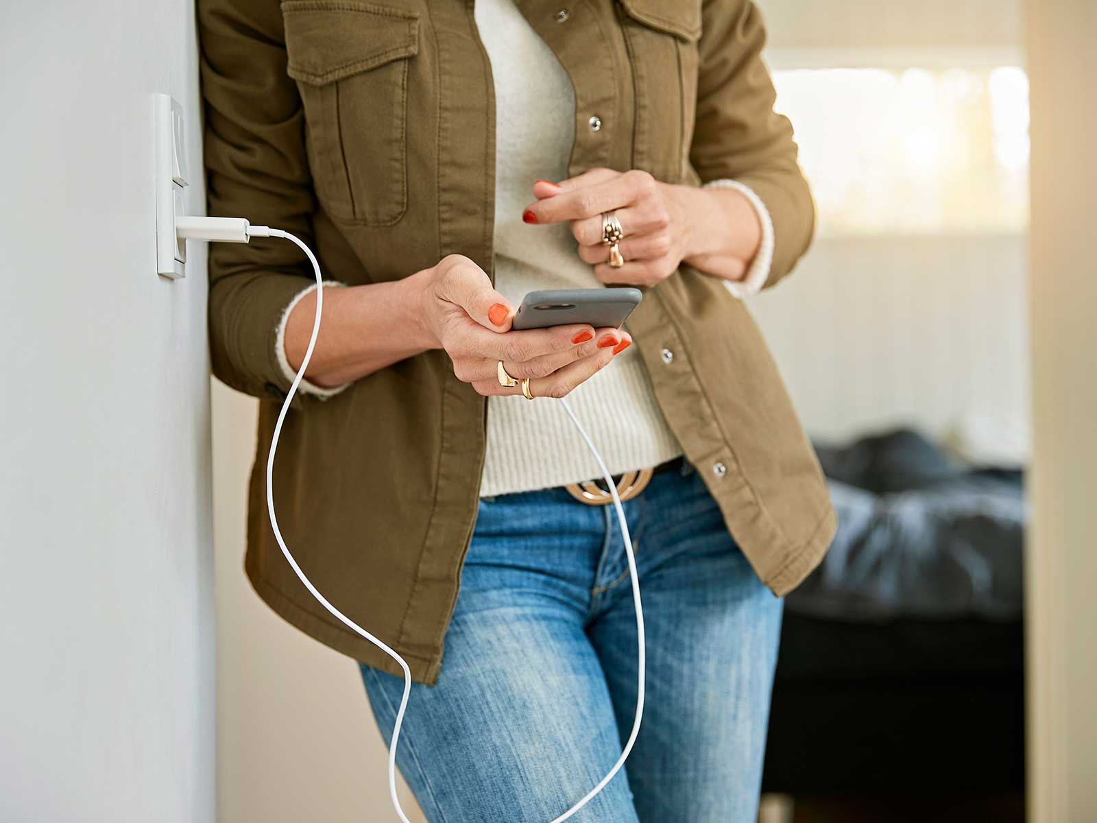 Kvinde holder en mobiltelefon i hænderne. Mobiltelefonen er sat til opladning i en stikkontakt på væggen.