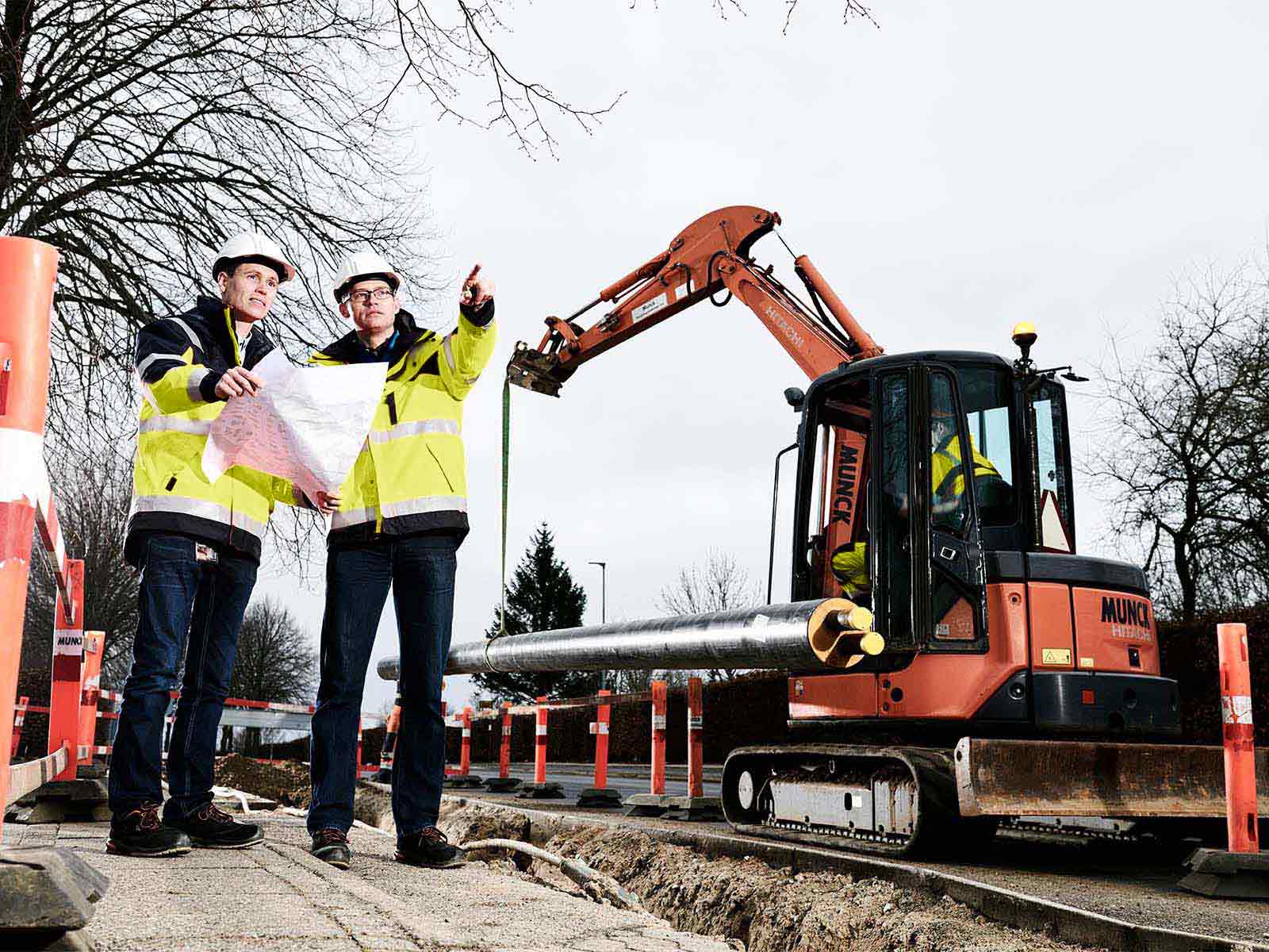 To projektledere står ved fjernvarmeprojekt med gravemaskine og fjernvarmerør i baggrunden