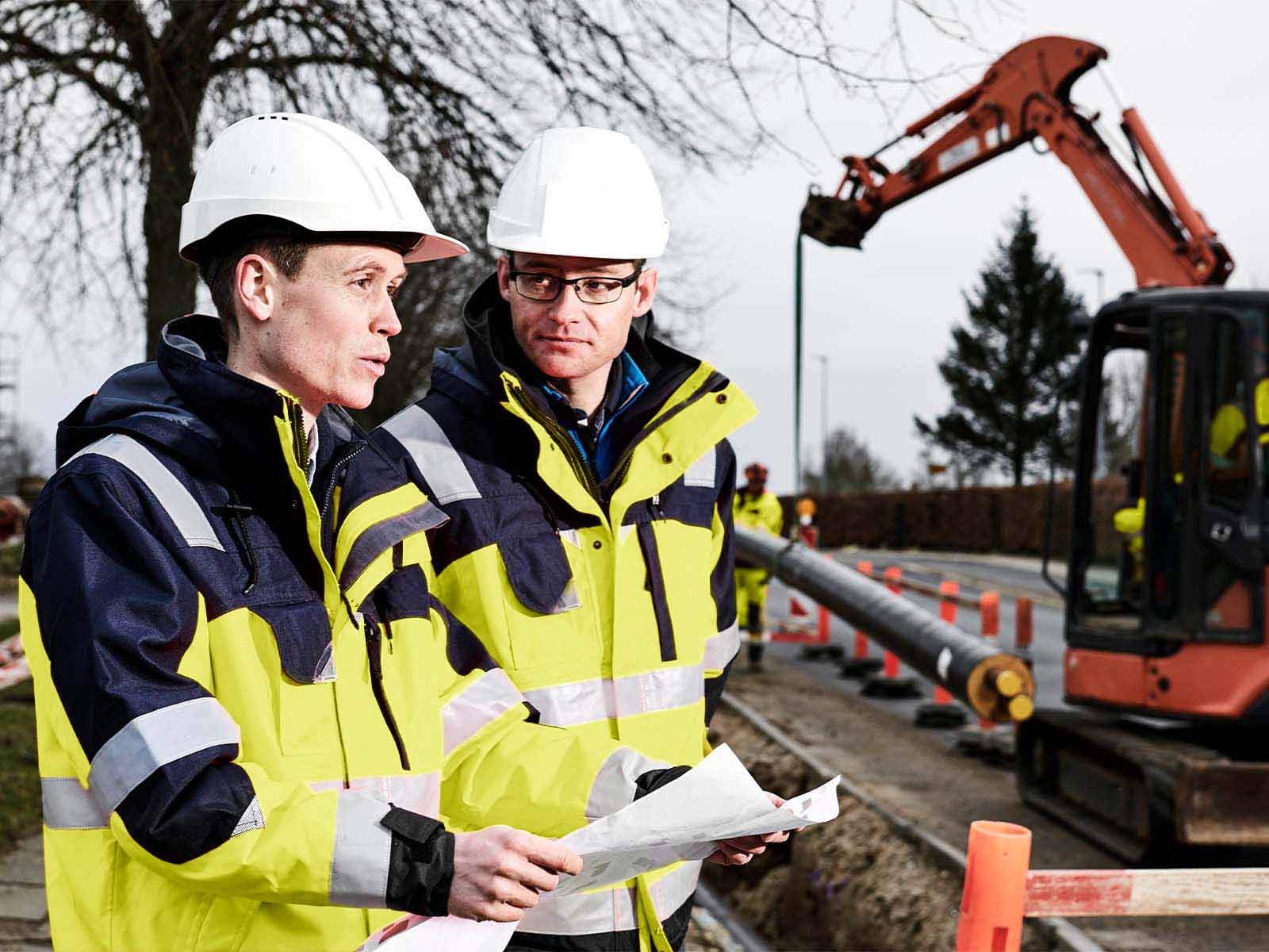 To projektledere står med kort ved fjernvarmeprojekt med gravemaskine i baggrunden