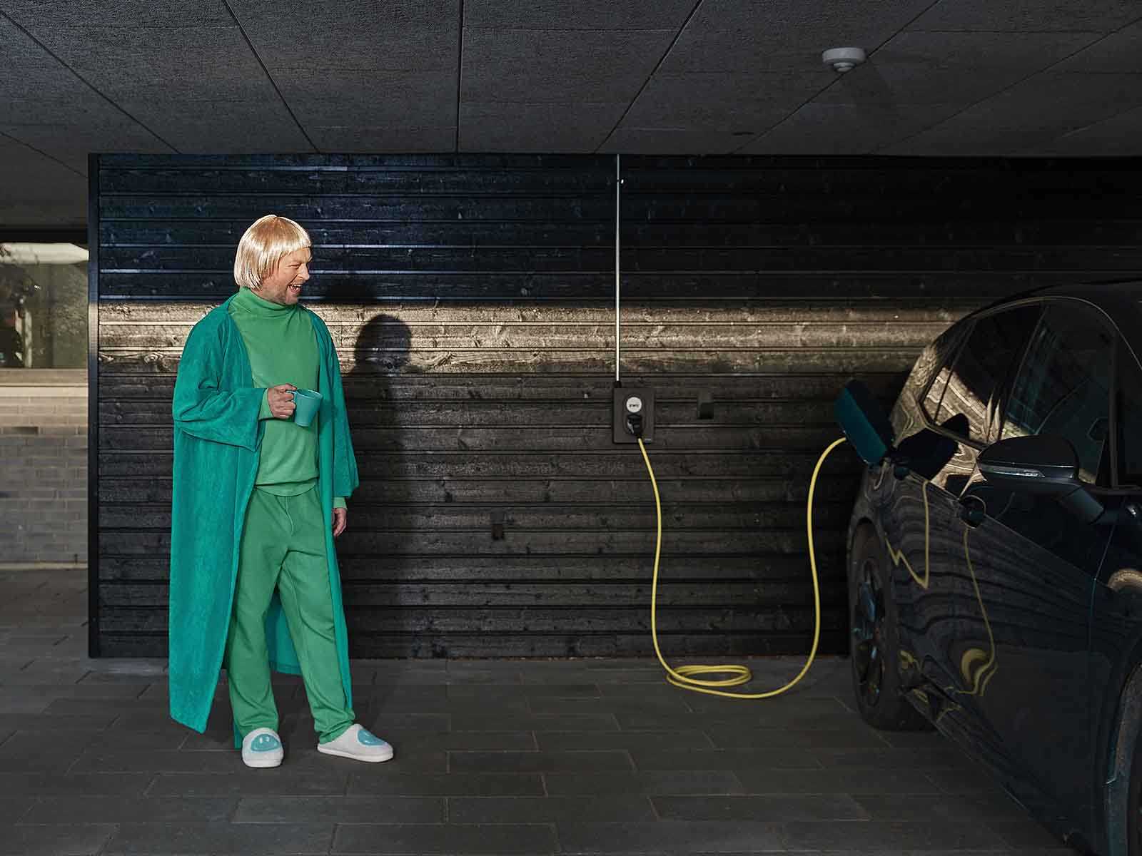 Mand i grøn morgenkåbe og paryk står i carport og beundrer sin elbil, der lader op med en ladeboks fra EWII. Kan et ladeabonnement betale sig? Eller er det bedre at betale for forbrug. Det undersøger vi her.