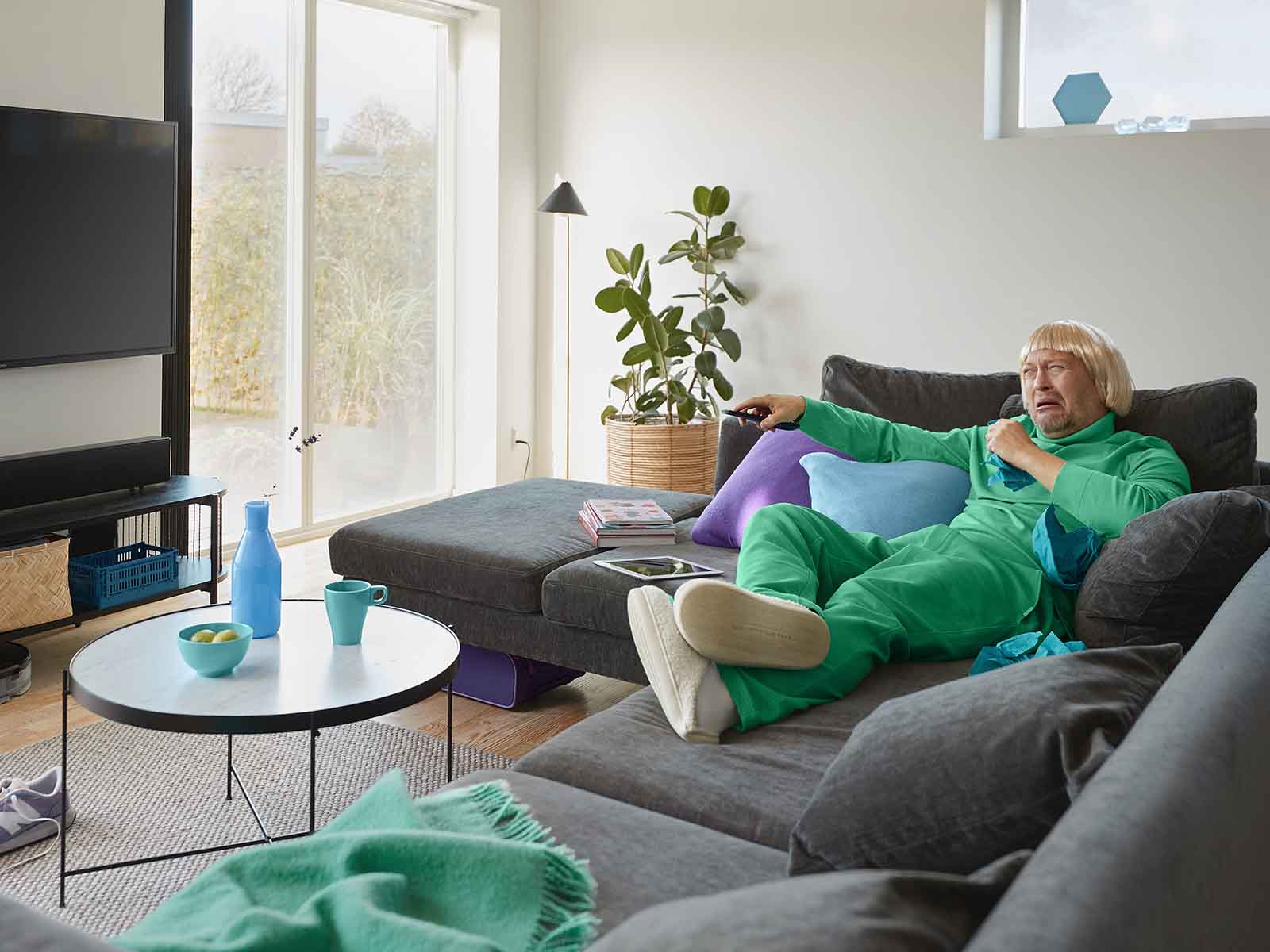 Mand i grønt tøj ligger på sofa og græder. Han streamer noget sørgeligt i fjernsynet.