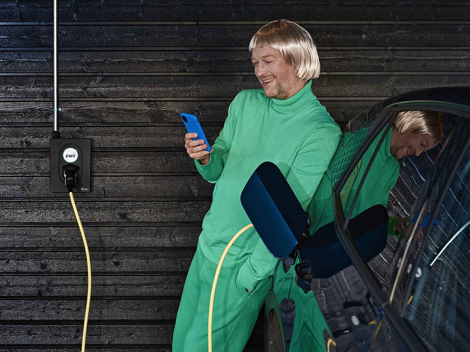 Mand læner sig op ad elbil med mobiltelefon i hånden. Han holder øje med elpriserne, så han har styr på, hvornår elprisen er lav, og det er billigst at lade.