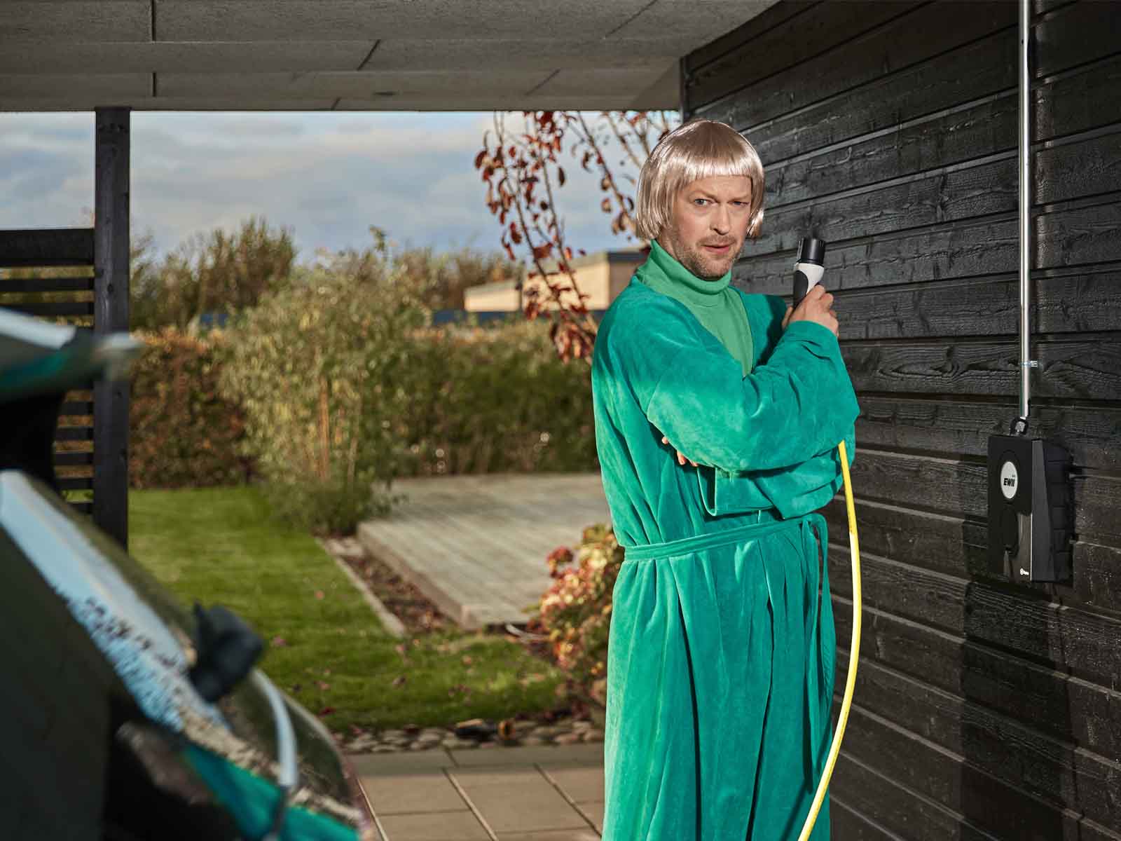 Mand i morgenkåbe og paryk står med ladekabel i carport, som var han James Bond, der holder en pistol. Hvor stort strømforbrug har din husstand? Se det typiske elforbrug for en gennemsnitsfamilie.