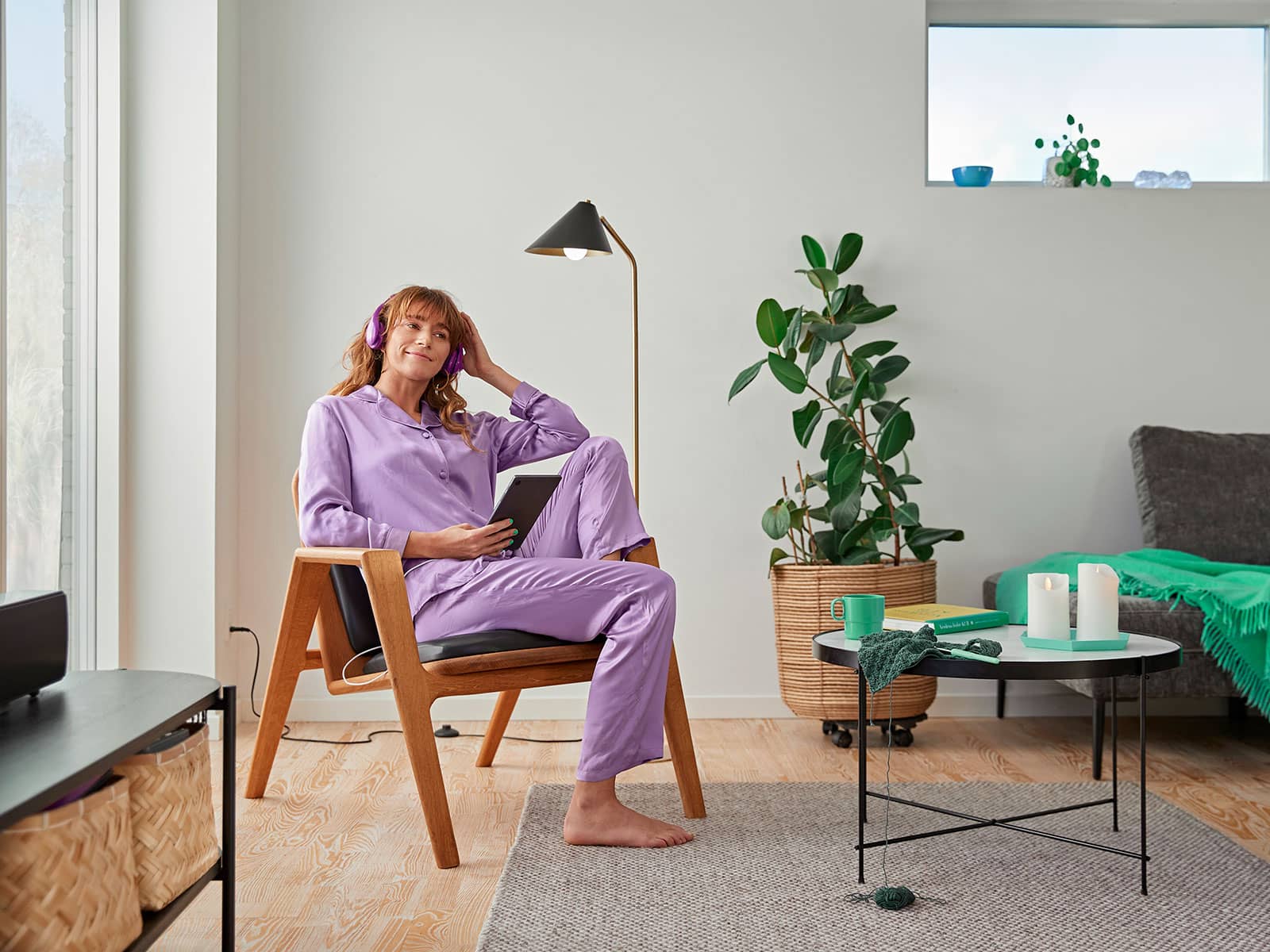 Kvinde i lilla pyjamas sidder i lænestol med sin tablet og lytter til en podcast. Hun har et lille elforbrug, så hun har valgt en Basis elaftale fra EWII.