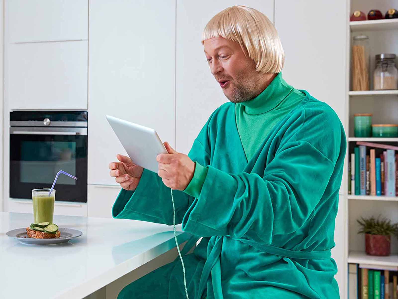 Mand i grønt morgenkåbe kigger på sin tablet i køkkenet. 
