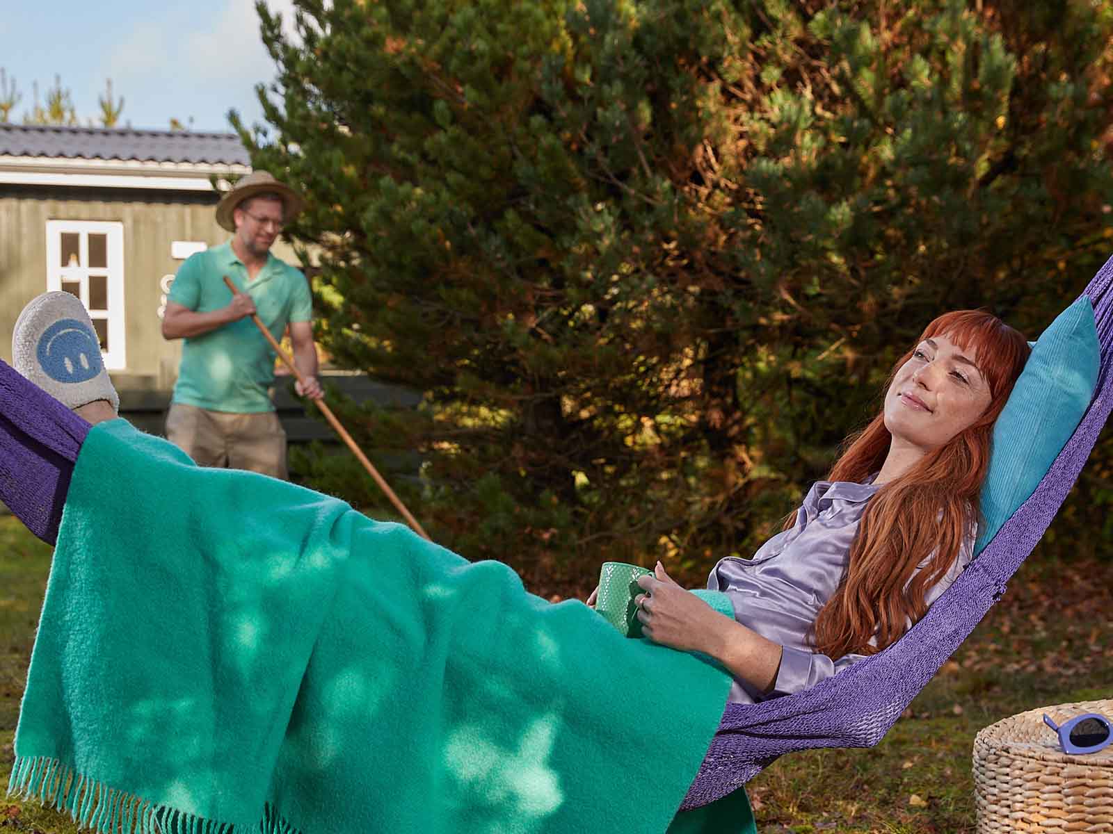 Ung kvinde i lilla pyjamas ligger i sin hængekøje og slapper af i sommerhuset mens hendes mand ordner haven.