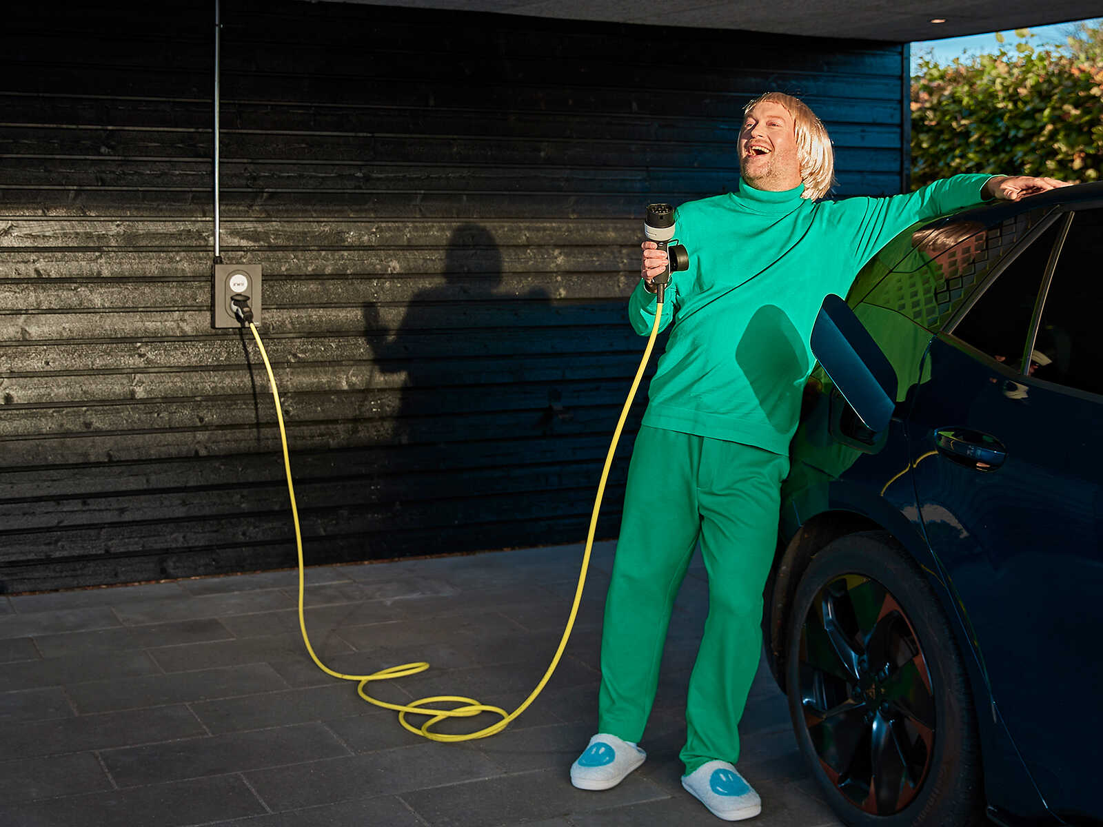 Glad mand læner sig afslappet op ad sin elbil, inden at sætter ladestikket i bilen.