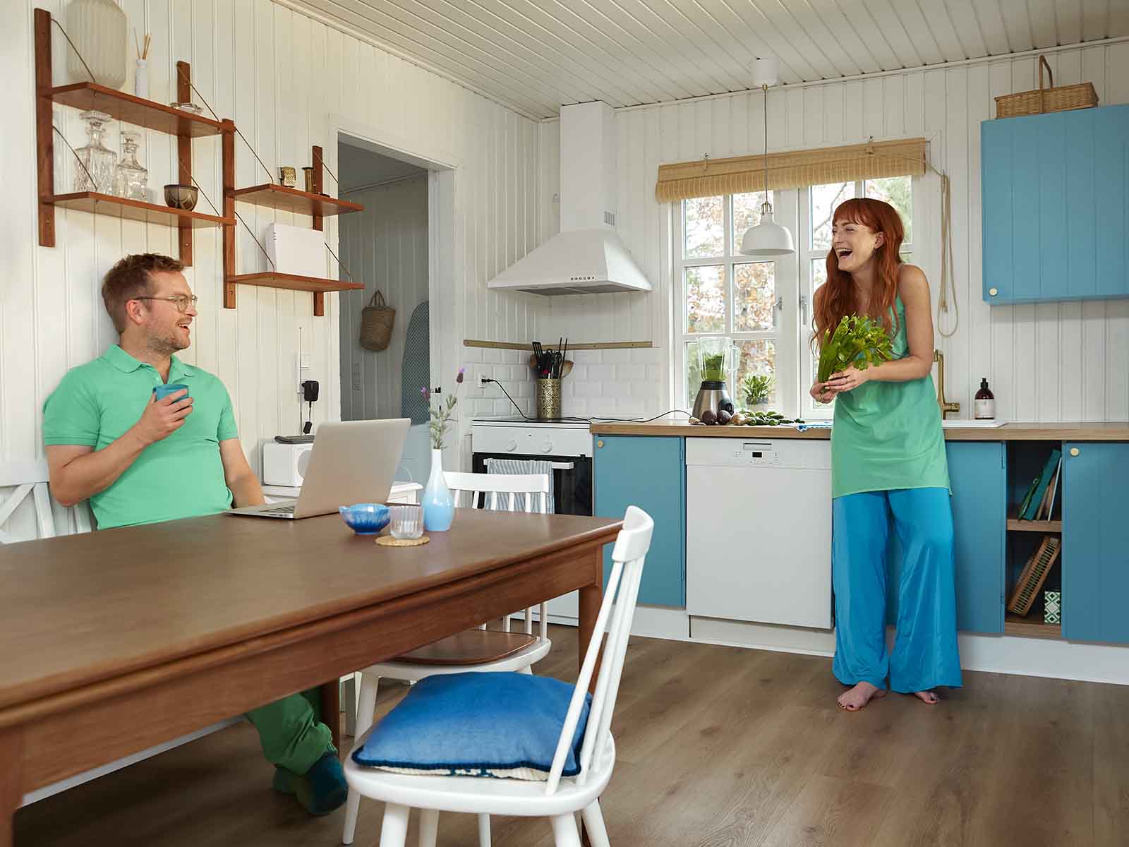 Mand sidder ved bord i køkken mens han smiler til kvinde der er gået i gang med at lave mad i køkkenet.