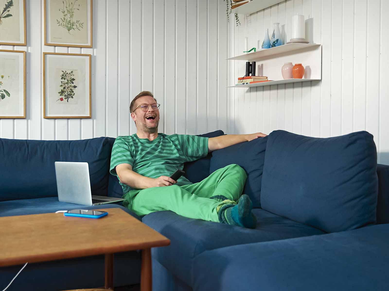 Mand i grønt tøj sidder i sin sofa i sommerhuset og griner fordi han ser noget sjovt i tv'et.