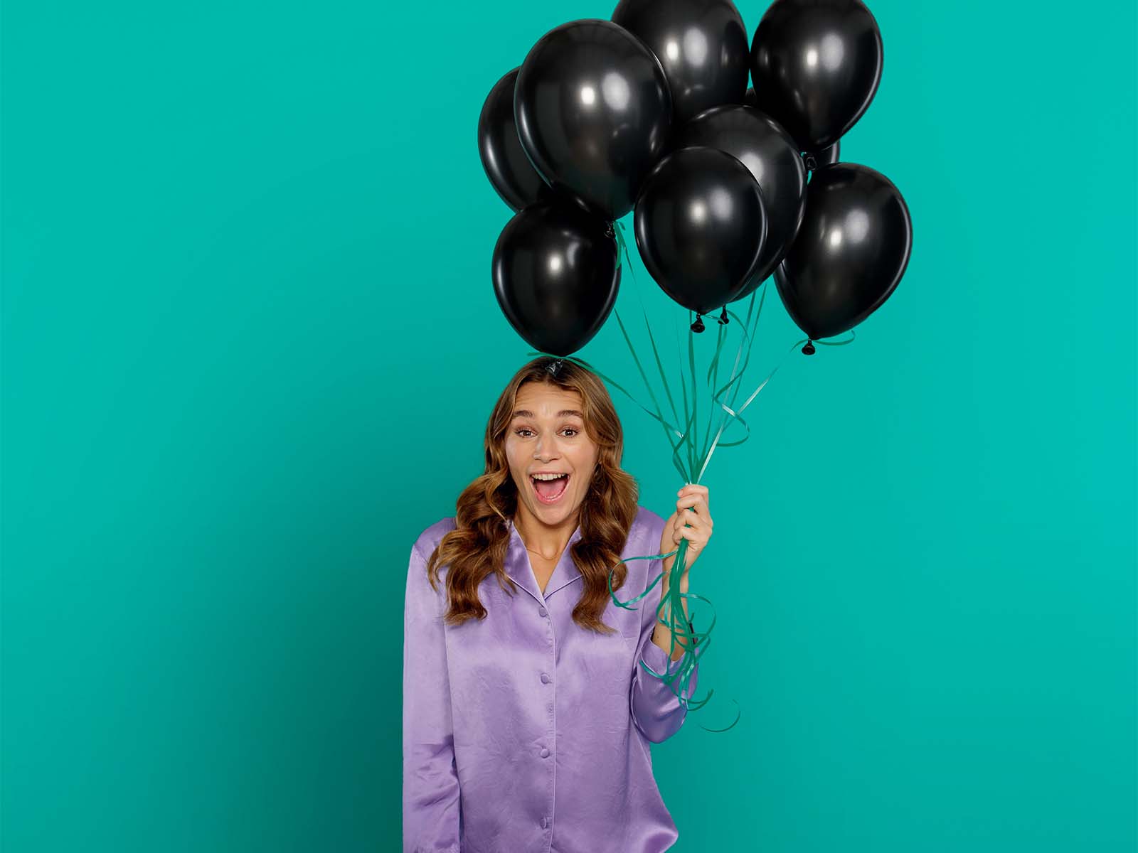 En kvinde i lilla pyjamas holder et bundt sorte balloner mens hun smiler stort og begejstret