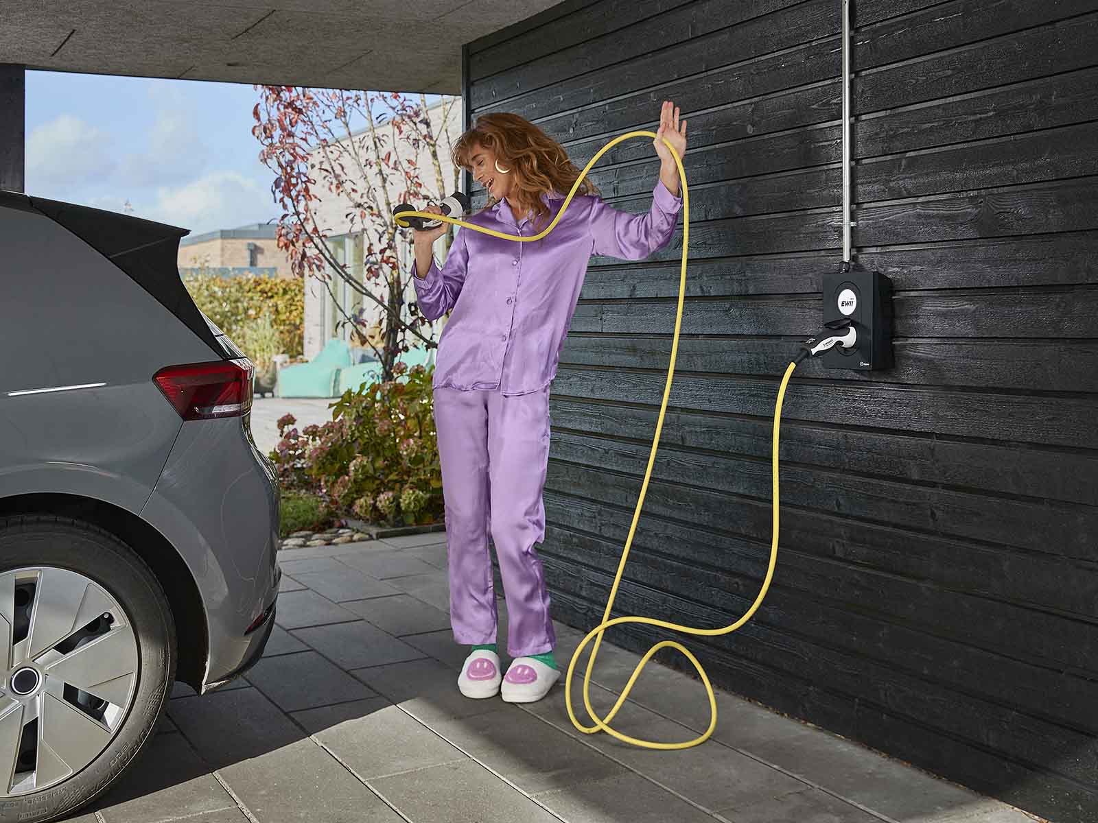 Kvinde i lilla pyjamas står i carport ved sin elbil og synger ind i ladekabel