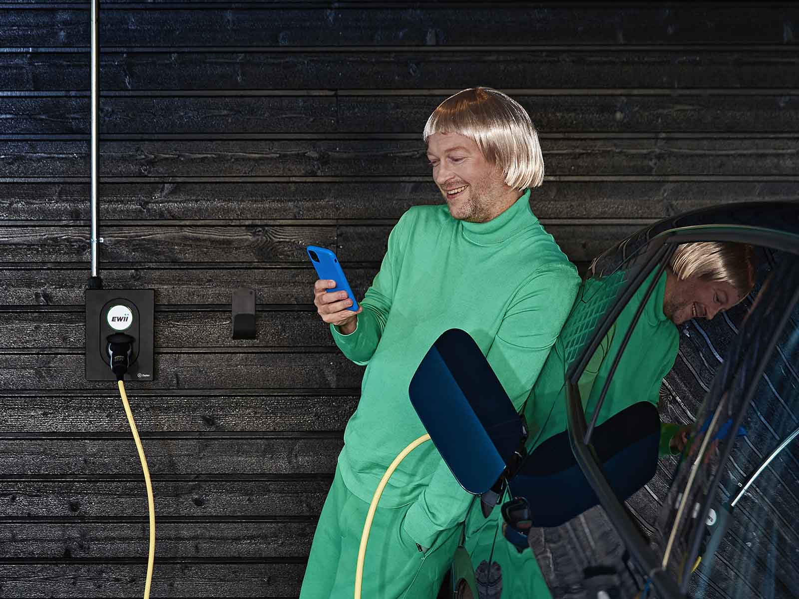 Mand læner sig afslappet op ad sin elbil med sin mobil i hånden, da han har en serviceaftale til sin ladeboks hos EWII. Han får fuld elafgiftsrefusion med serviceaftalen.