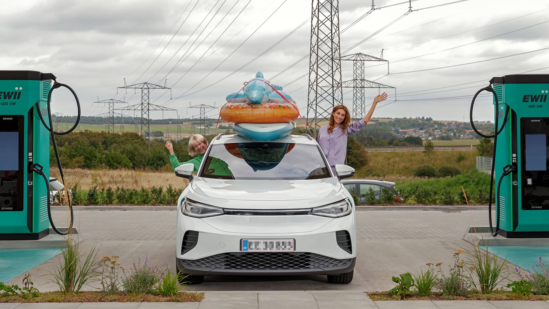 Mand i grønt tøj og kvinde i lilla pyjamas i bil med badedyr på taget lader ved lynladestation på vej på sommerferie i elbil