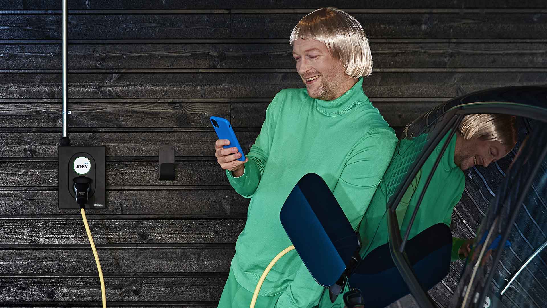 mand tjekker mobiltelefon imens han lader sin elbil op derhjemme