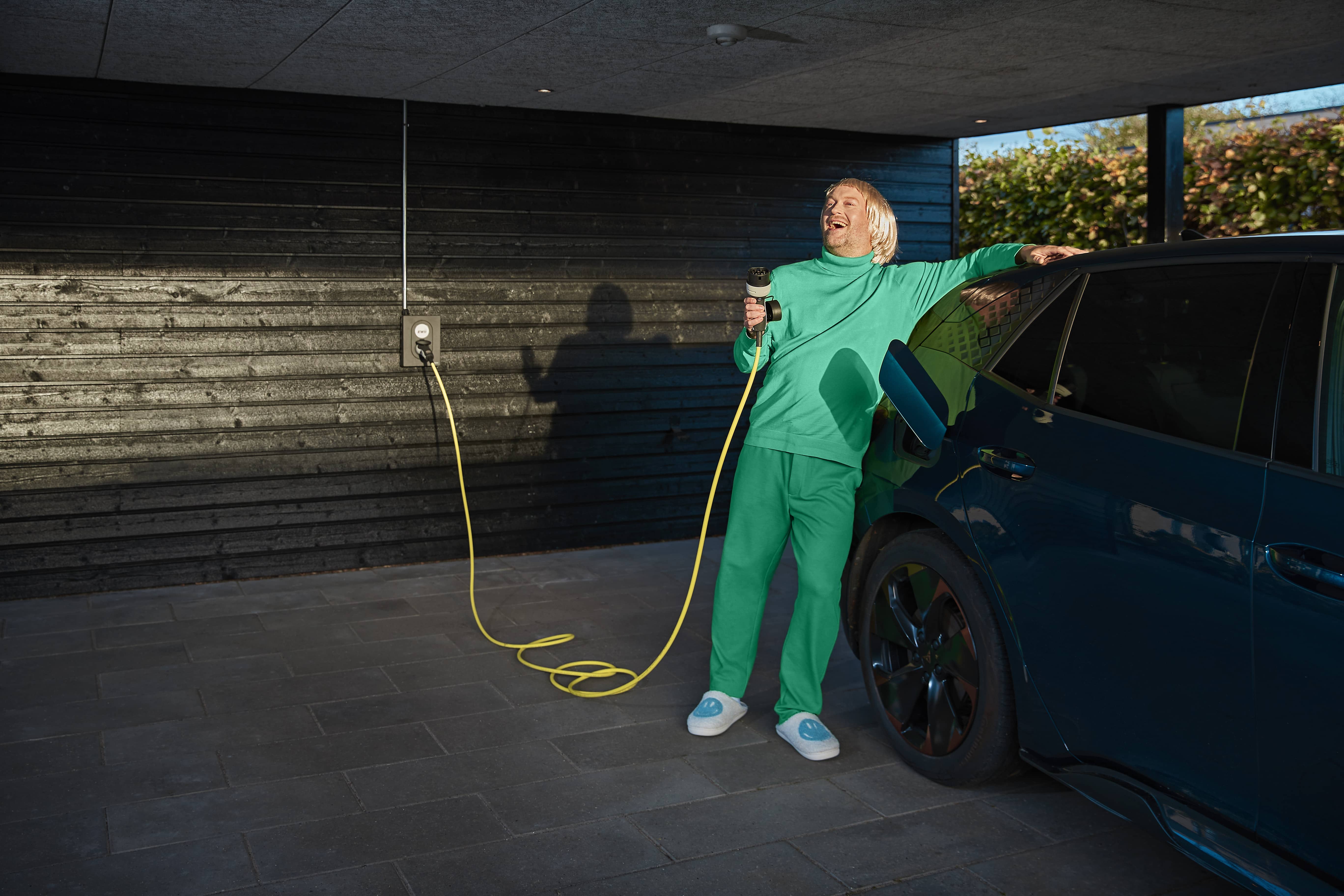 Glad mand læner sig op af sin elbil, inden han sætter stikket i sin bil.