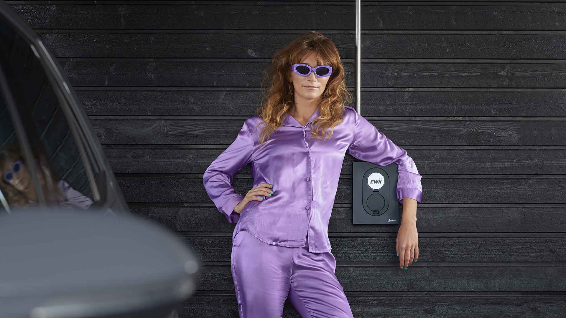 kvinde med solbriller og pyjamas læner sig op af sin ladeboks i carporten