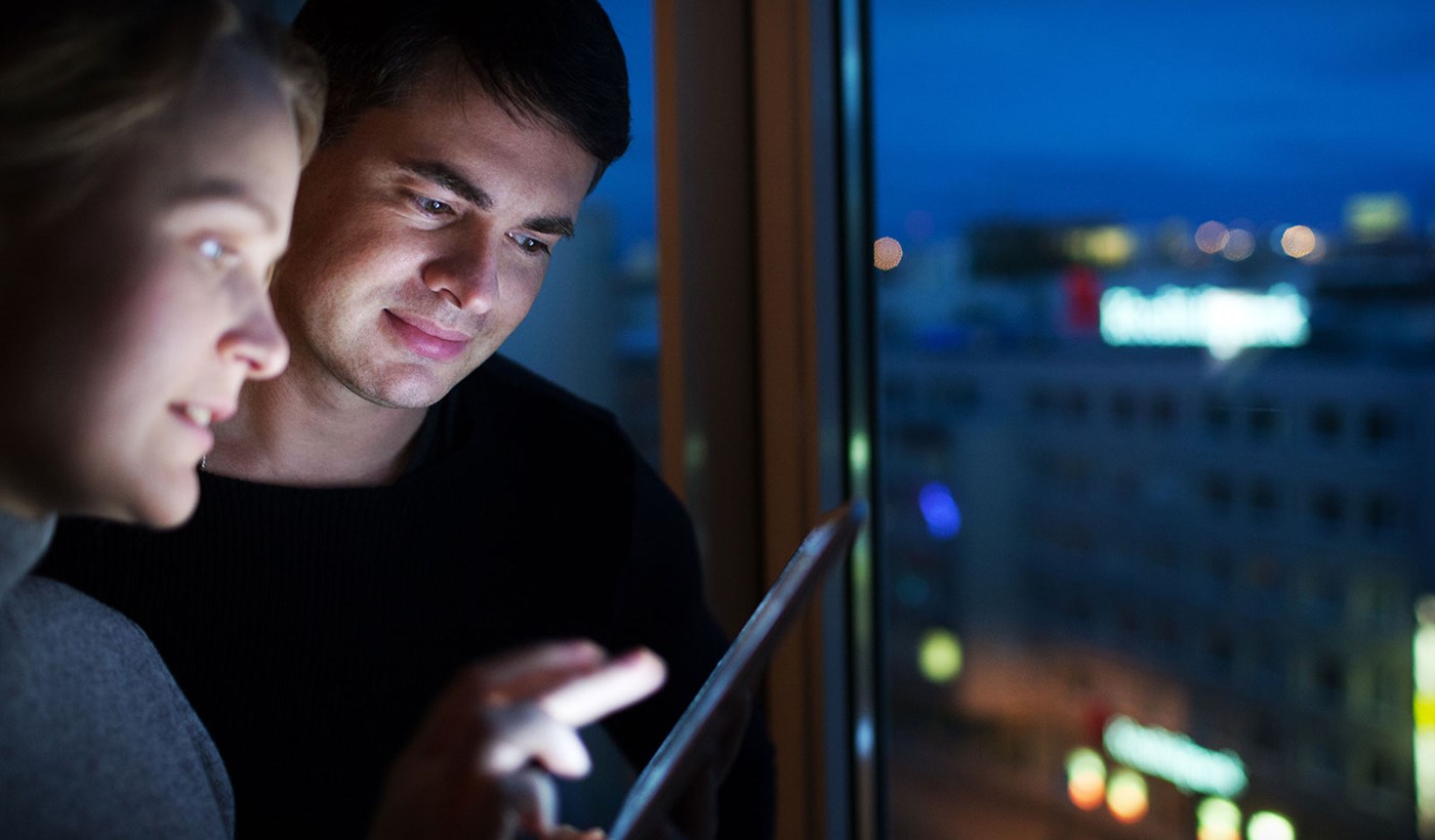 Ungt par kigger på iPad ved vindue om aftenen, hvor der typisk er billigere strøm end om dagen. 