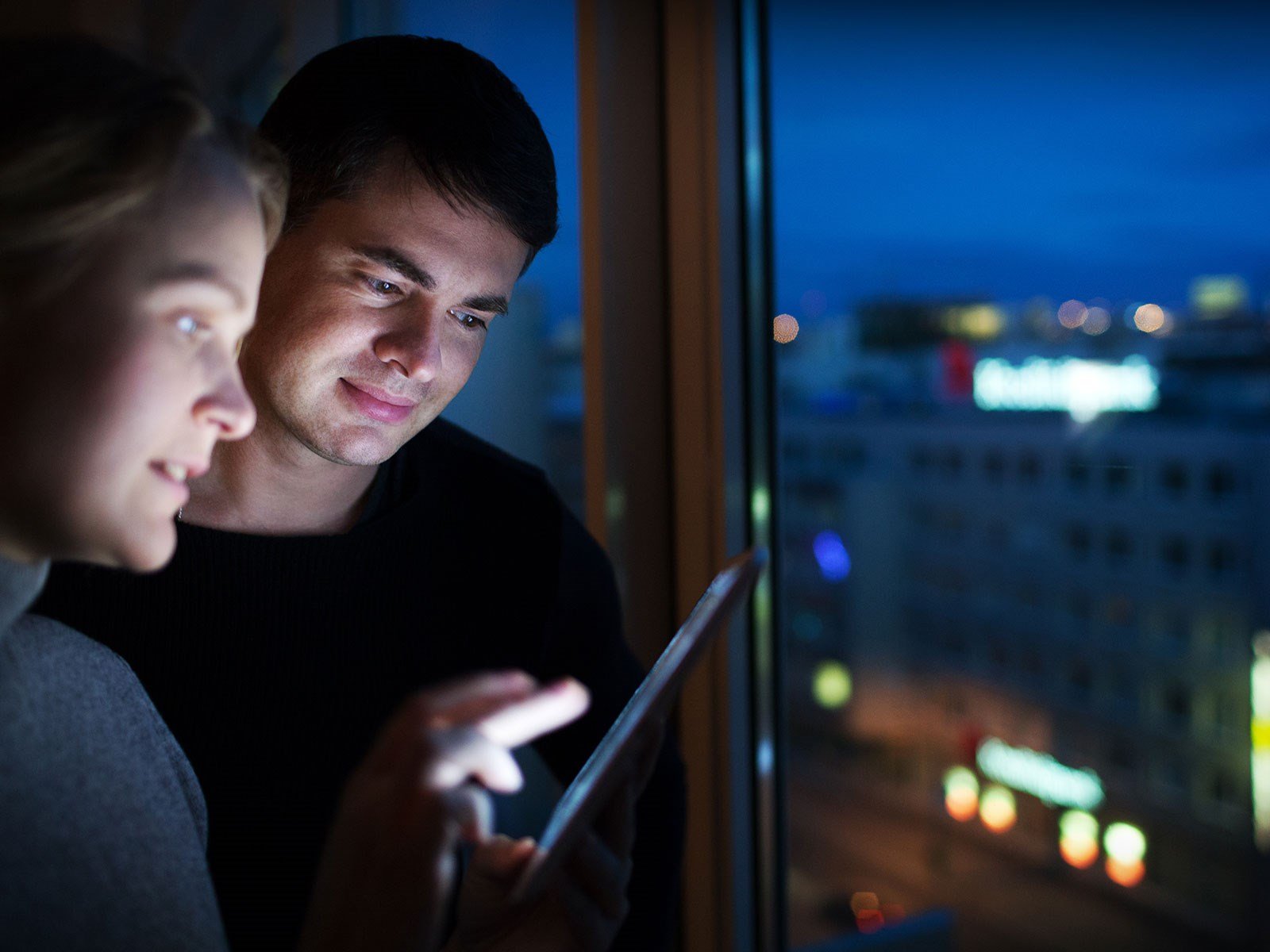 Ungt par kigger på iPad ved vindue om aftenen, hvor der typisk er billigere strøm end om dagen. 